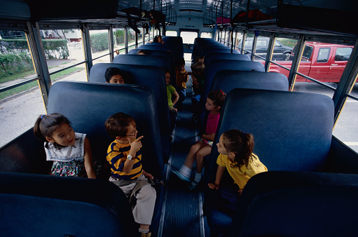 Diverse elementary school-aged children seated singly on a school bus talk to their classmates across the aisle.