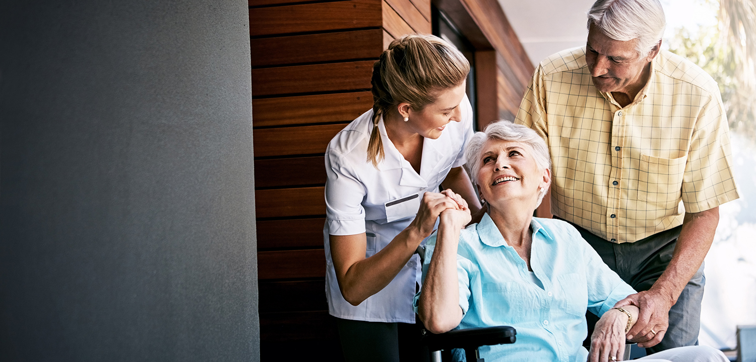 elderly woman in wheelchair smiling and thanking her caregiver while an elderly smiling man stands next to her