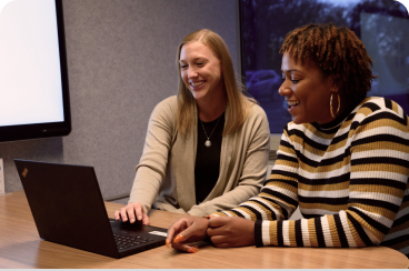 Two women sit at a desk and converse while one uses a laptop.