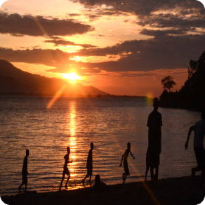 People run along the shore as an organge-reddish sun set reflects on the water.